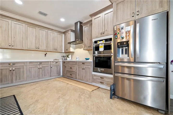 Kitchen featuring stainless steel appliances, light countertops, wall chimney exhaust hood, light brown cabinetry, and recessed lighting