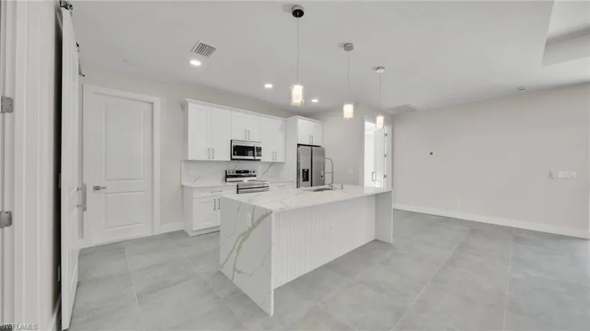 Kitchen with light stone counters, hanging light fixtures, white cabinets, appliances with stainless steel finishes, and an island with sink