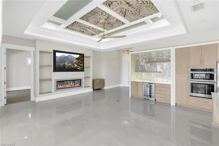Unfurnished living room with built in shelves, a tray ceiling, beverage cooler, a glass covered fireplace, and a ceiling fan