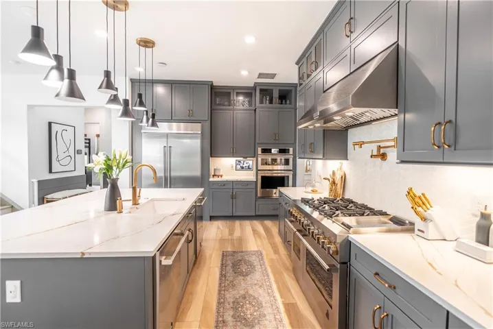 Gourmet kitchen featuring a large central island with an undermount sink and golden-toned faucet, light wood-finish flooring, full-height cabinetry with golden-toned hardware, stainless steel appliances, and a gas range with an overhead hood
