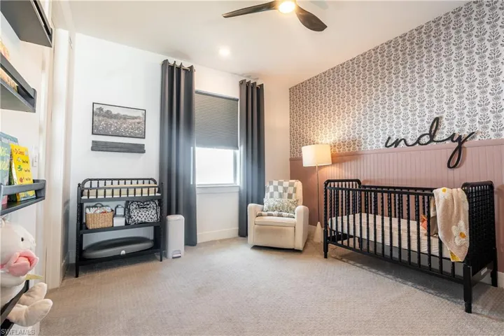 Carpeted room featuring a ceiling fan with integrated lighting, a window with roller shade and dark curtains, built-in shelving, and a decorative accent wall with wainscoting