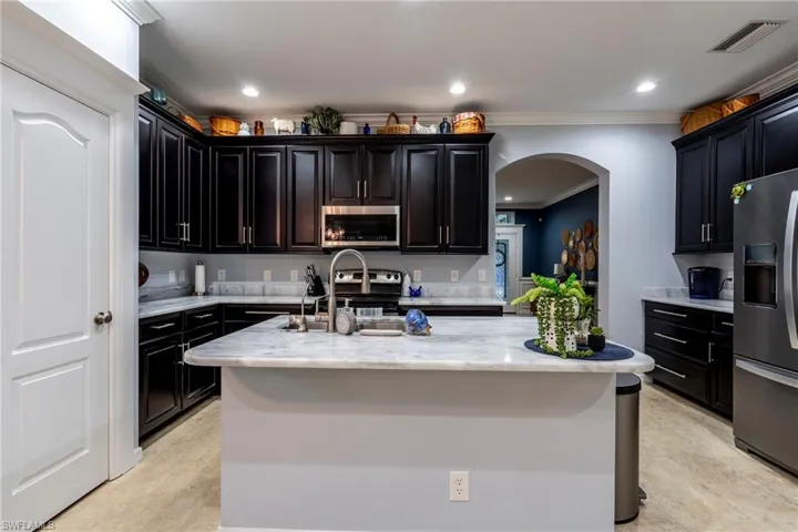 Kitchen featuring dark cabinetry, crown molding, appliances with stainless steel finishes, arched walkways, and light stone counters