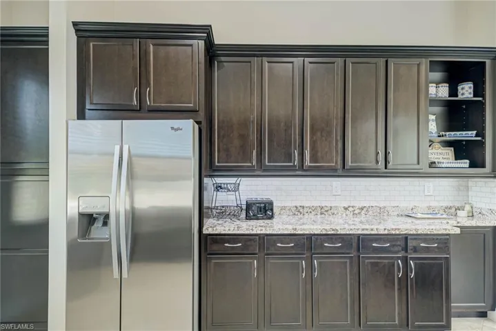 Kitchen with open shelves, stainless steel fridge with ice dispenser, backsplash, light stone counters, and dark brown cabinetry