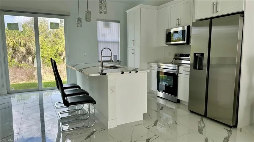 Kitchen featuring stainless steel appliances, light marble finish flooring, an island with sink, pendant lighting, and white cabinetry