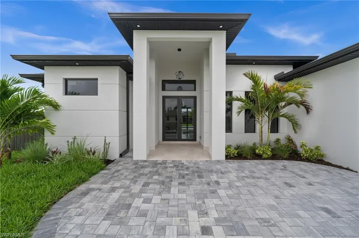 Contemporary exterior featuring a recessed entry, dark-framed double glass doors, a paver driveway, white stucco finish, and dark fascia with integrated lighting