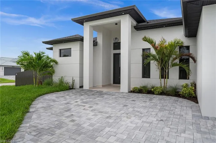 Contemporary stucco exterior featuring a grand entryway, dark frame windows, a paver driveway, and mature palm trees