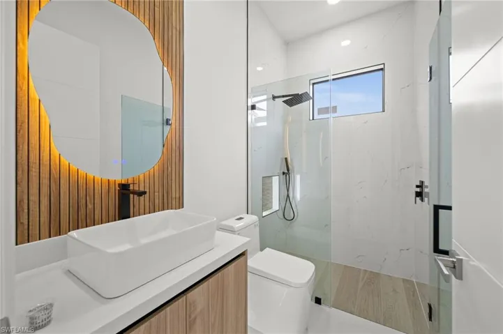Modern bathroom featuring a custom-shaped backlit mirror, slatted wood-finish accent wall, vessel sink, and a glass-enclosed shower with a rainfall showerhead