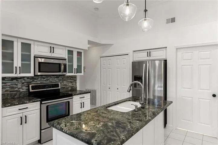 Kitchen featuring stainless steel appliances, dark stone countertops, white cabinets, decorative light fixtures, and a center island with sink