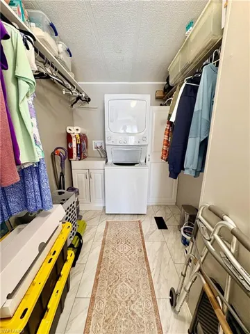 Laundry area featuring a textured ceiling, stacked washer and clothes dryer, light marble finish flooring, and cabinet space