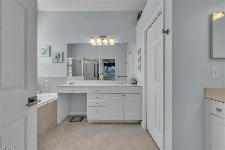 Bathroom featuring a white vanity with drawers and integrated seating, a full-width wall mirror, four-light vanity fixture, drop-in soaking tub with tile surround, and tile flooring
