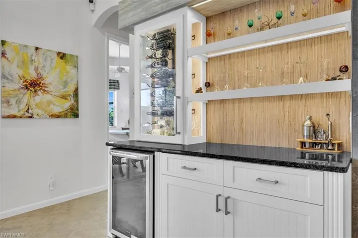 Built-in bar featuring custom white cabinetry, a glass-front wine refrigerator, dark stone-finish countertops, and wood-paneled backsplash with integrated floating shelving