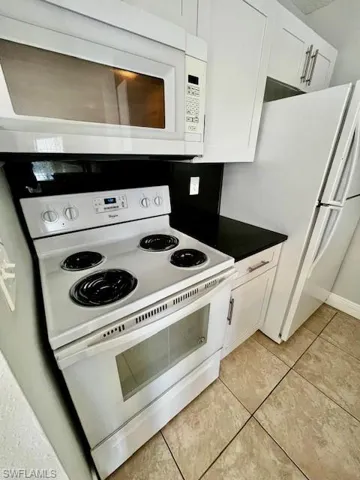 Kitchen with light tile patterned floors, dark countertops, white cabinetry, and white appliances