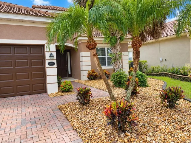 Welcoming front elevation featuring a two-car garage with wood-finish doors, clay tile roofing, and a professionally landscaped entry with mature palm trees, decorative stone hardscaping, and colorful tropical plantings including flowering shrubs and ornamental foliage