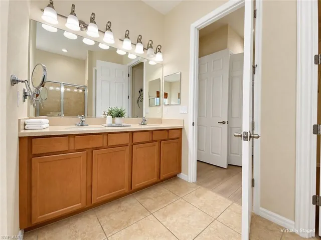 Dual-vanity master bathroom featuring warm wood-finish cabinetry, ceramic tile flooring, and a large framed mirror with integrated vanity lighting