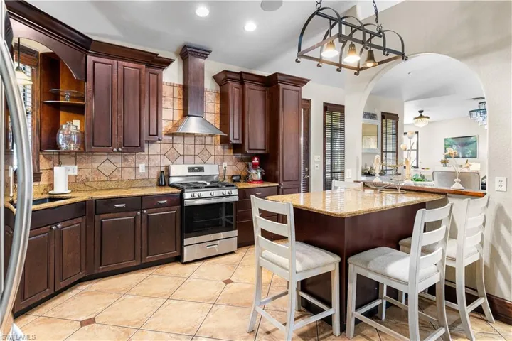 Kitchen with arched walkways, dark wood finish cabinets, light stone countertops, stainless steel appliances, and a kitchen bar