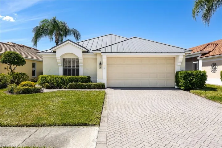 Stucco exterior featuring a metal roof, two-car garage, paver driveway, and mature landscaping