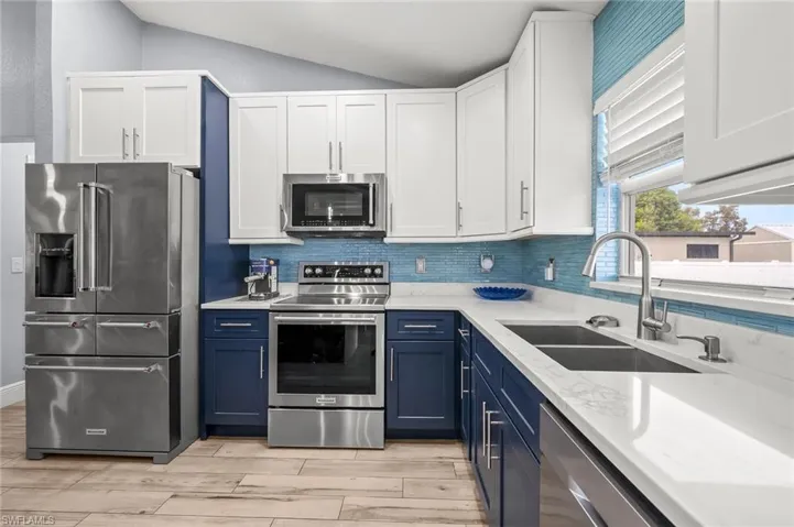 Two tone kitchen with two tone cabinets, stainless steel appliances, light stone counters, vaulted ceiling, and light wood finished floors
