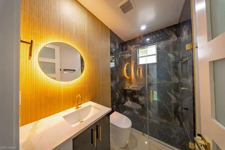 Contemporary bathroom featuring a back-lit oval mirror, a vanity with a white countertop and rectangular undermount sink, a gold-finish faucet, and a walk-in shower with clear glass enclosure and black-patterned wall tiles