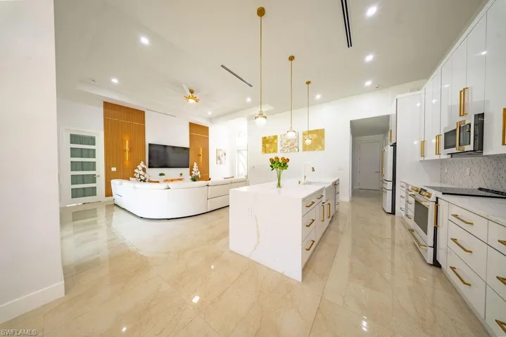 Open-concept interior featuring large-format tile flooring, a white kitchen with gold-toned hardware, an oversized kitchen island with pendant lighting, recessed ceiling lights, and a white paneled door with frosted glass inserts
