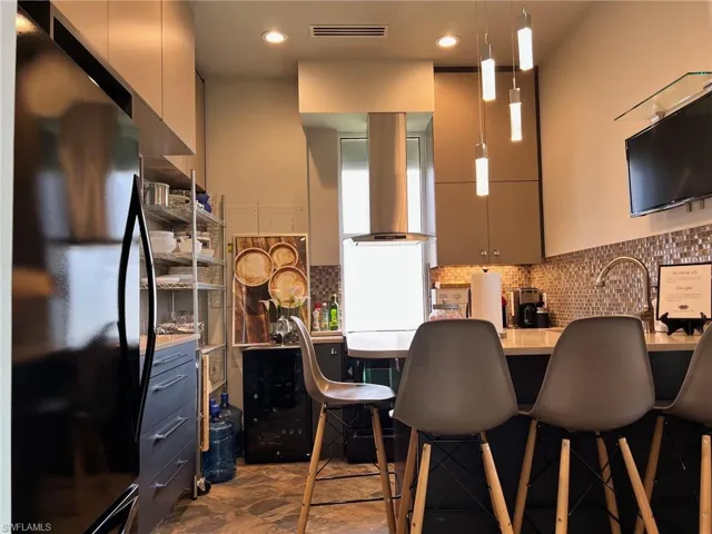 Kitchen featuring freestanding refrigerator, wall chimney range hood, a breakfast bar, decorative backsplash, and hanging light fixtures