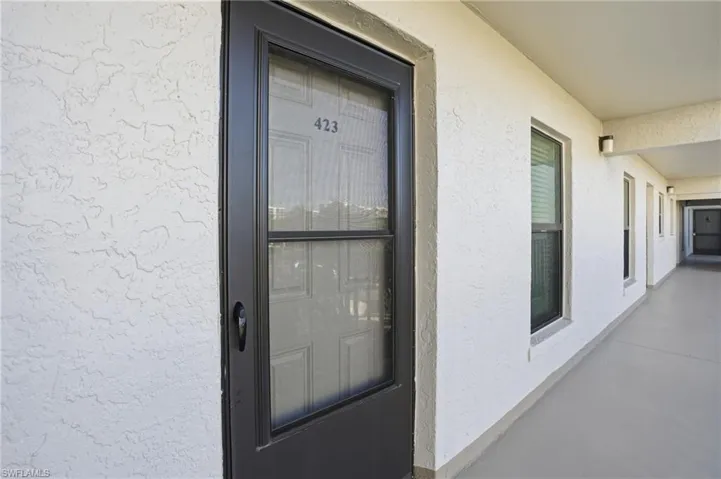 Unit entrance featuring a dark-framed screen door with house number 423, a textured white stucco exterior, and a covered outdoor corridor with multiple windows and exterior lighting fixtures - Virtually Edited Image