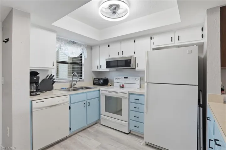Galley kitchen featuring white appliances, a double basin stainless steel sink, wood-finish flooring, white upper cabinetry, and light blue lower cabinetry - Virtually Edited Image