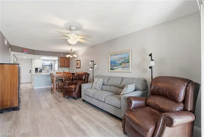 Open concept living area featuring wood-finish flooring, a ceiling fan with integrated lighting, and a partial wall separating a kitchen with light wood cabinetry and a breakfast bar - Virtually Edited Image