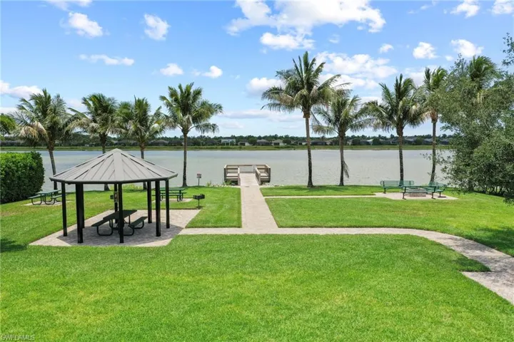 Expansive green space featuring a lakeside gazebo with picnic tables, a concrete pathway, a wooden dock, and mature palm trees