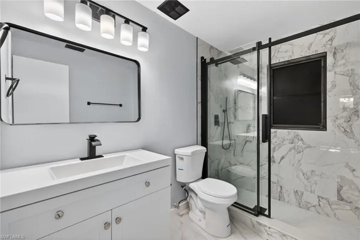 White vanity with integrated rectangular sink, stone-finish shower surround, matte black hardware, glass-enclosed shower, and four-light vanity fixture