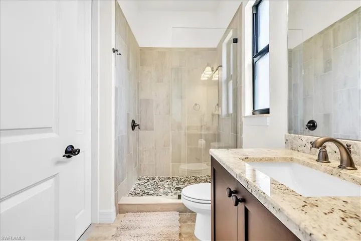 Bathroom featuring a glass-enclosed shower with pebble flooring and tiled walls, a dark wood-finish vanity with a granite countertop, an undermount sink, and a bronze-finish faucet