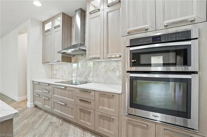 Kitchen with stainless steel double oven, wall chimney exhaust hood, light wood-style flooring, light brown cabinets, and light countertops