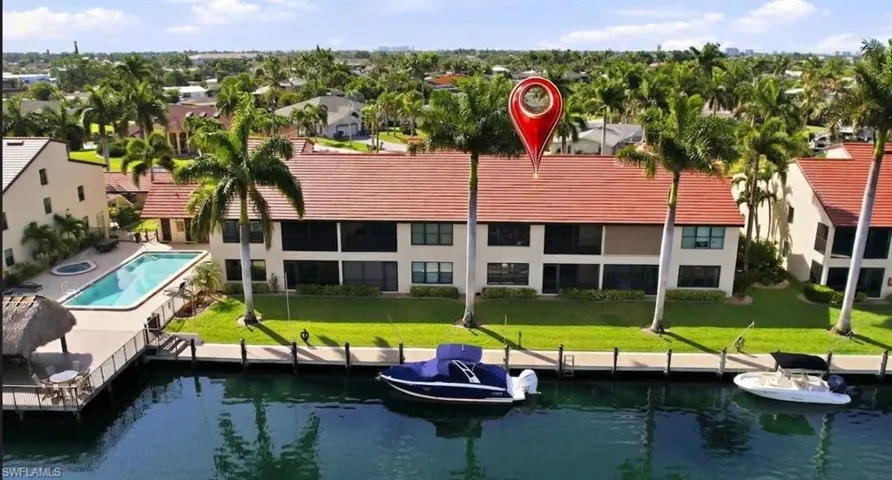 Waterfront condominium building with red tile roofing, featuring private boat docks, a swimming pool, and a thatched-roof cabana