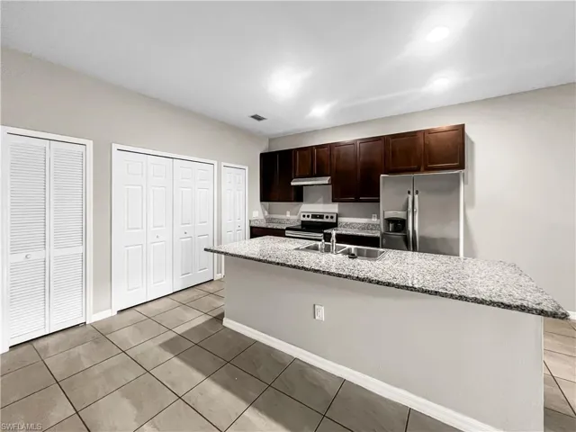 Kitchen featuring a granite-look countertop island with an undermount sink, dark wood-finish cabinetry, stainless steel appliances, and tile flooring