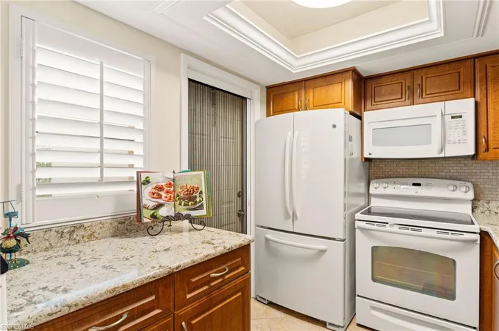 Kitchen featuring light Granite countertops, white appliances, decorative backsplash, brown cabinetry, and a raised ceiling