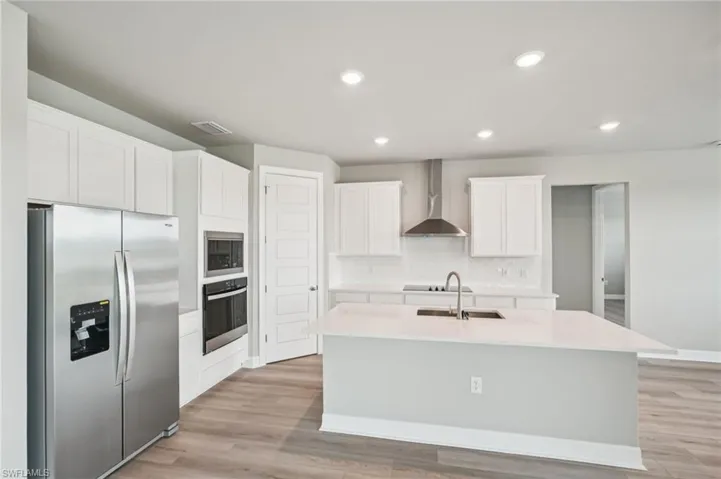 Kitchen featuring black appliances, wall chimney exhaust hood, a sink, a center island with sink, and white cabinetry