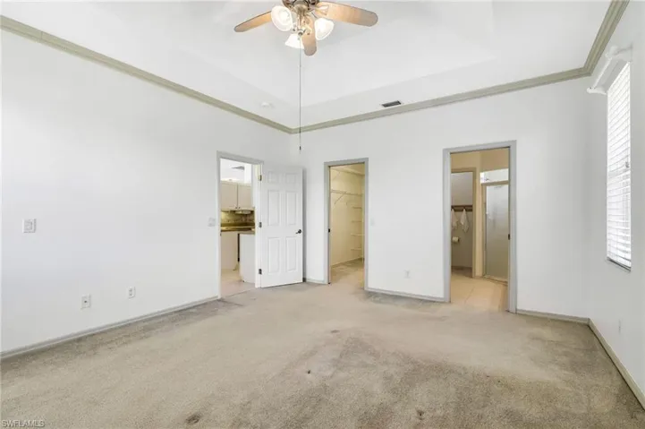 Unfurnished Primary bedroom with light colored carpet, a walk in closet, ornamental molding, ensuite bathroom, and a ceiling fan