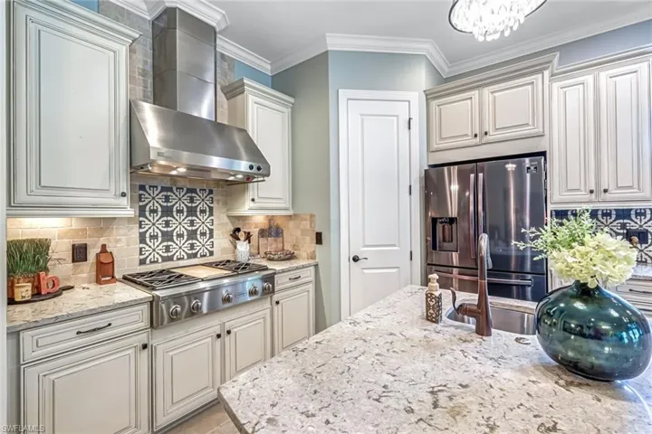 Kitchen with wall chimney exhaust hood, stainless steel appliances, tasteful backsplash, light stone countertops, and a chandelier