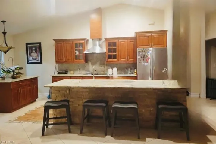 Kitchen with brown cabinets, stainless steel fridge, tasteful backsplash, wall chimney exhaust hood, and high vaulted ceiling