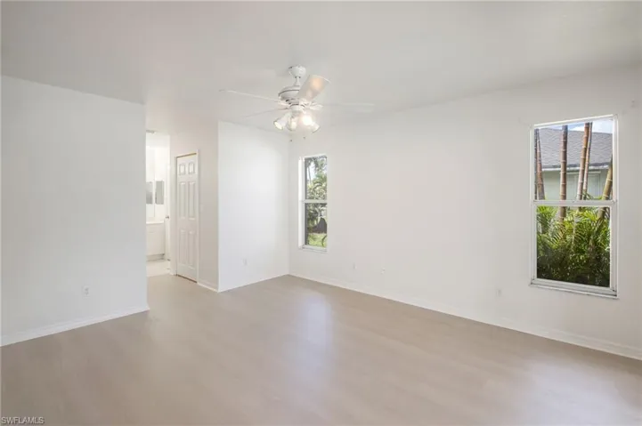 Spacious room featuring light wood-finish flooring, a white ceiling fan with integrated lighting, multiple windows providing natural illumination, and crisp white walls