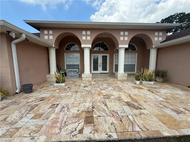 Spacious stone-tiled patio featuring a stucco exterior with arched entryway, decorative columns, and double entry doors