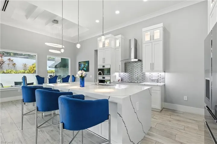 Kitchen with wall chimney exhaust hood, crown molding, tasteful backsplash, a center island with sink, and light stone countertops