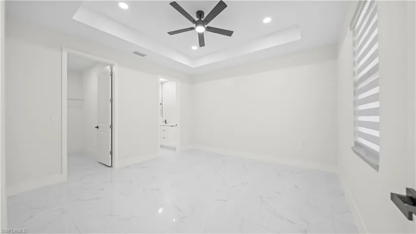 Unfurnished bedroom with a tray ceiling, a walk in closet, recessed lighting, a ceiling fan, and light marble finish floors