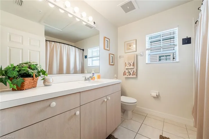 Bathroom featuring a single vanity with light wood-finish cabinetry, a large vanity mirror with strip lighting, a window with blinds, and tiled flooring