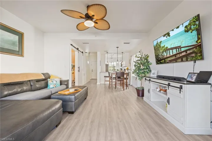 Open-concept living space featuring light wood-finish flooring, a ceiling fan with rattan-style blades, and a white entertainment console with barn door hardware