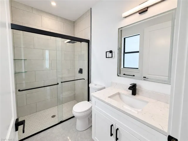 Bathroom featuring a vanity with a white countertop and rectangular undermount sink, matte black hardware, glass-enclosed walk-in shower with tile surround, and a frameless mirror with mounted light fixture