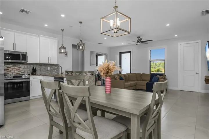 Open concept living area featuring a kitchen with white cabinetry, stainless steel appliances, and a tiled backsplash, leading into a dining space with a light-colored table and chairs, and a living area with a ceiling fan and a window providing natural light