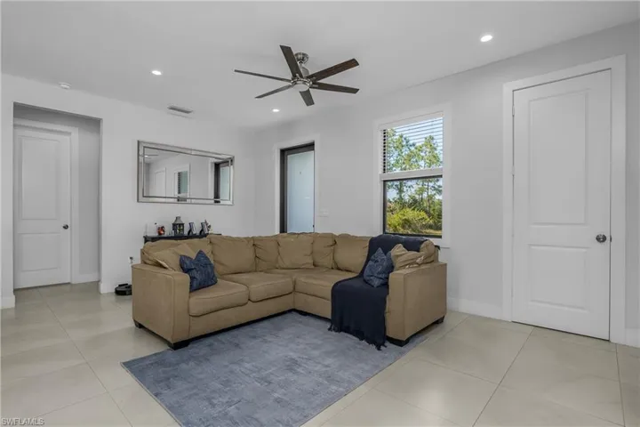 This bright living area features light-colored tile flooring, a ceiling fan, recessed lighting, and a window with blinds offering views of outdoor greenery