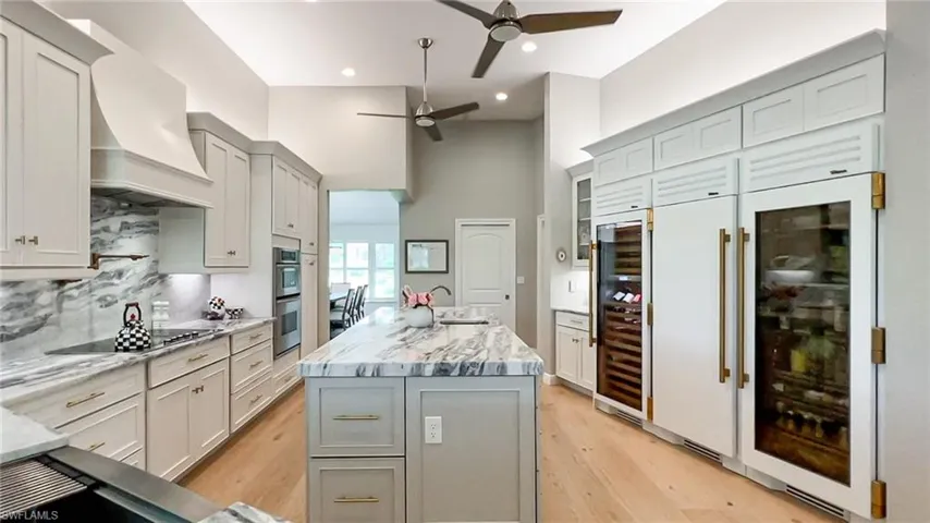 Kitchen with light stone countertops, ceiling fan, wine cooler, light wood-style floors, and an island with sink