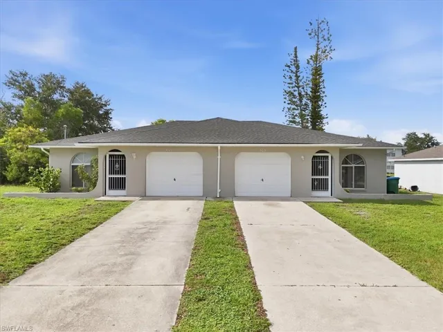 Duplex property featuring a neutral stucco exterior, dark shingle roof, arched windows, covered entryways with security doors, and individual garage access