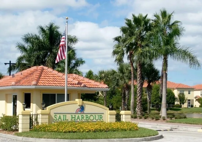 Entry monument with "SAIL HARBOUR" signage, framed by established palm trees and low-lying yellow flowering plants